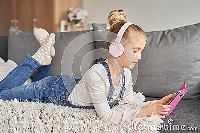 Girl laying on couch in headphones, listening to music with her smarthphone. Little girl watching cartoons on her phone. Listening Stock Photo