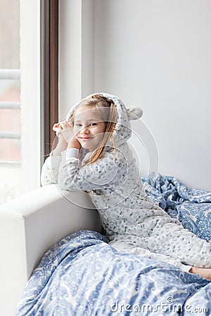 A girl in light gray pajamas sits on a bed by the window, the child woke up in the morning, morning yoga Stock Photo