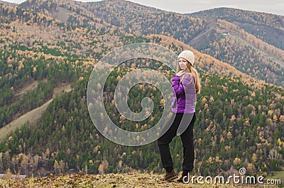 A girl in a lilac jacket looks out into the distance on a mountain, a view of the mountains and an autumnal forest by an overcast Stock Photo