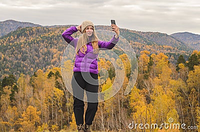 A girl in a lilac jacket makes a salfi on a mountain, a view of the mountains and an autumnal forest by a cloudy day Stock Photo