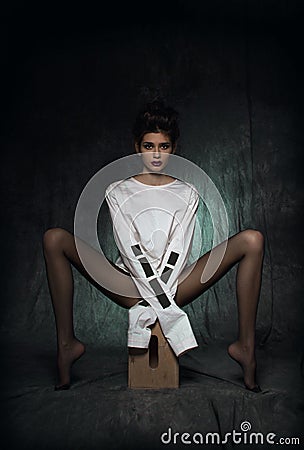 A girl with long legs in black tights and a white straitjacket sits on a wooden pedestal with her legs spread to the side Stock Photo