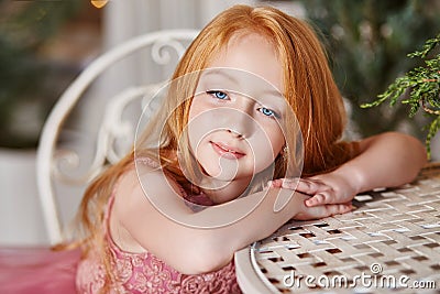 Girl with long red hair in a pink dress sitting at the table. Carnival holiday birthday. Portrait of a cheerful red-haired girl Stock Photo