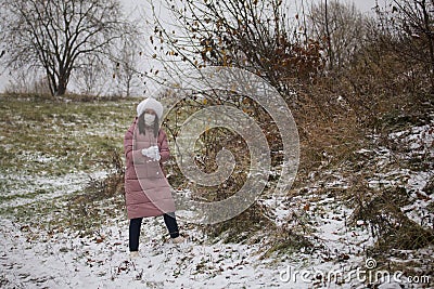 A girl in a medical mask and winter clothes on a walk in the park. Holds a ball of snow in his hand. The ground is covered with th Stock Photo