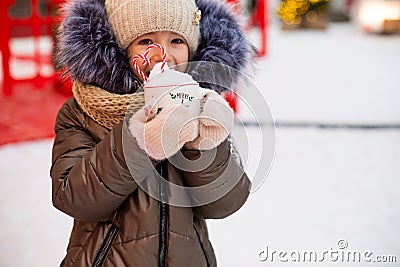 Girl with mug with snow, candy cane and inscription Merry and Bright in her hands outdoor in warm clothes in winter at festive Stock Photo