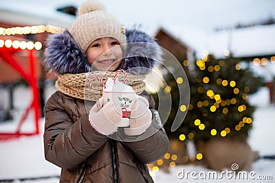 Girl with mug with snow, candy cane and inscription Merry and Bright in her hands outdoor in warm clothes in winter at festive Stock Photo