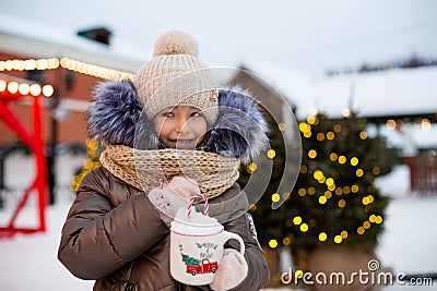 Girl with mug with snow, candy cane and inscription Merry and Bright in her hands outdoor in warm clothes in winter at festive Stock Photo