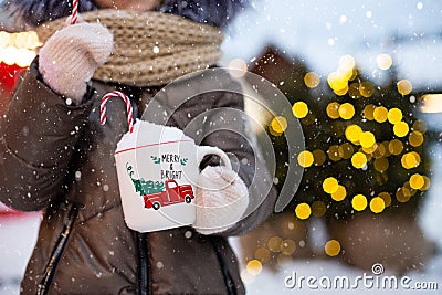 Girl with mug with snow, candy cane and inscription Merry and Bright in her hands outdoor in warm clothes in winter at festive Stock Photo