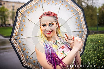 A pretty girl in a beautiful dress with bright multi-colored braids and shiny makeup is whirling in a spring park in Stock Photo