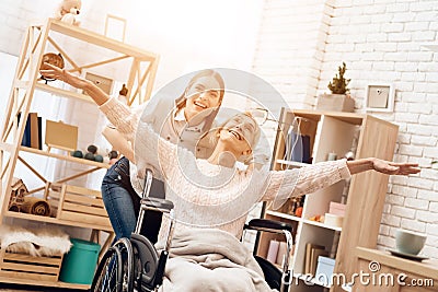 Girl is nursing elderly woman at home. Girl is riding woman in wheelchair. Woman feels like flying. Stock Photo