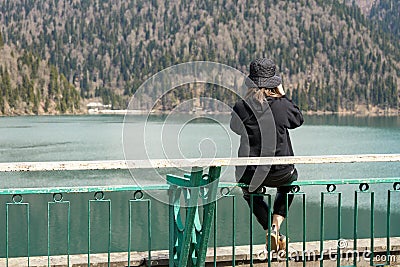 A girl in a panama hat on her head sits on a fence high above the pond and takes pictures of herself on her phone against the back Stock Photo