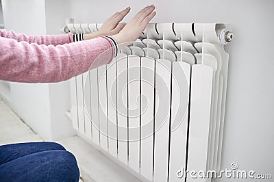 Girl in a pink sweater and blue jeans warms hands near a radiator Stock Photo