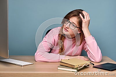 A girl in a pink sweater sits at a table with a computer and books, and, holding on to her head, looks exhaustedly at Stock Photo