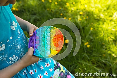 Girl plays with touch anti-stress silicone pop it toy outdoor in the garden. Fingers press on rainbow soft bubbles and buttons. Stock Photo