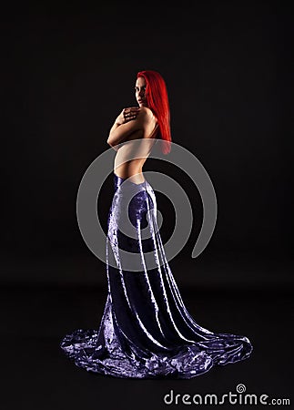 A girl with red hair, in a long lilac skirt, poses against a dark background in a contoured light Stock Photo
