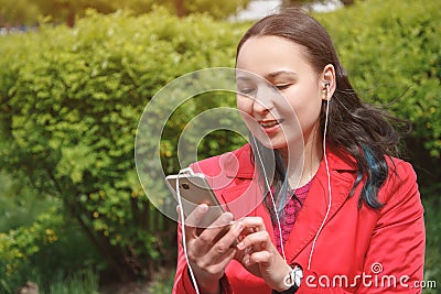 Girl in a red jacket in the Park listening to music with headphones earbuds with a smartphone in hand Stock Photo