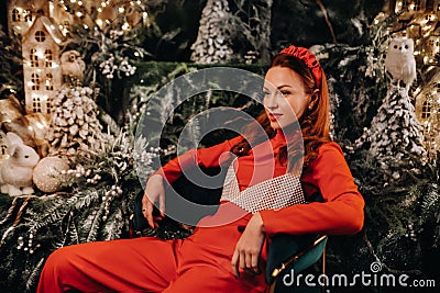 A girl in a red suit is sitting on a chair near the Christmas background. a woman in red on the background of Christmas trees and Stock Photo