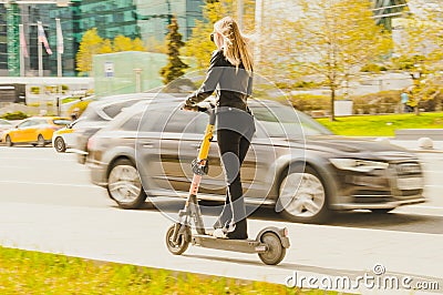 girl rides a kick scooter around the city along the road. Rear view of woman in black casual clothes riding electric scooter Editorial Stock Photo