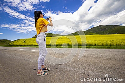 Girl on the road, in a yellow T-shirt, taking a photo in a flowering field, against the backdrop of mountains Stock Photo