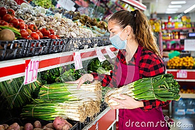 Girl salesperson in a protective mask puts leeks on a display case Stock Photo