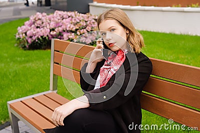 A girl sits on a bench in the street and speaks on the phone, orders a taxi by phone. Girl holds mobile in hands and laughs, Stock Photo