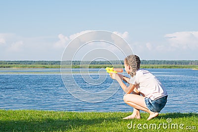 The girl is sitting on the green grass and playing with a water pistol, against the blue sky and the lake, there is a place for an Stock Photo