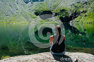 A girl sitting on a rock, looking at a spectacular view of the mountain reflecting in the lake. Stock Photo