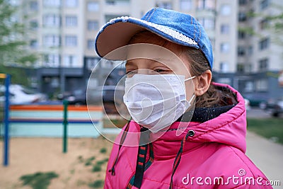 A girl sitting on a seesaw on playground near high-rise buildings with apartments, a medical mask on her face protects against Stock Photo