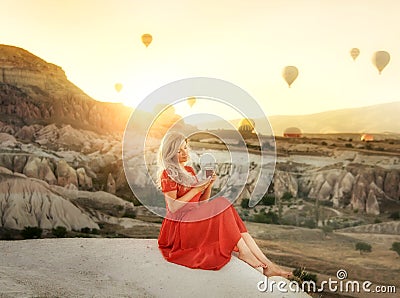 A girl sitting on the top of a cliff with a glass of Turkish tea at dawn with a view of the mountains of Cappadocia and balloons i Stock Photo