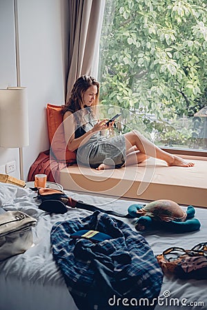 Woman tourist sitting near window using mobile phone in room with mess in hotel Stock Photo