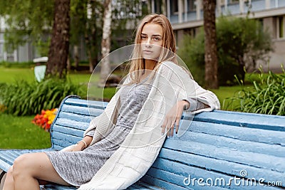 A girl in stylish clothes and beautiful looks is sitting on a blue bench in a summer park. Stock Photo