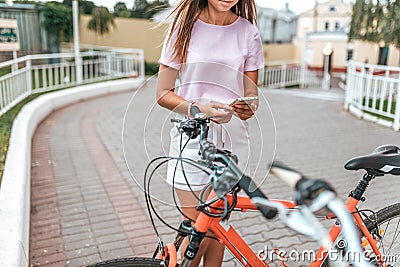 Girl summer stands bicycle, activates application buying rental bicycle in parking lot, in hands of phone. Online Stock Photo