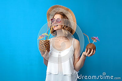 Girl in a sun hat and glasses drinks tropical cocktails made of pineapple and coconut on a blue isolated background, a woman with Stock Photo