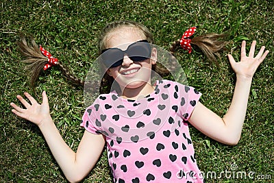 Girl in sunglasses lie on green grass, dressed in pink clothes with hearts, bright sun, summer outdoor, top view Stock Photo