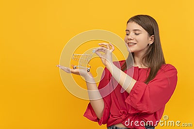 Girl to red blouse holds grocery cart on her hand. Portrait of young woman with shopping cart on the palm of her hand on yellow Stock Photo