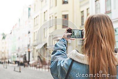 Girl travels in the old town and makes a photo landscape on a smartphone. Back view. Tourist in a jeans jacket photographs a city Stock Photo