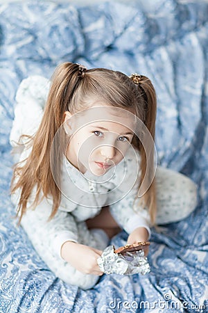 A girl with two ponytails in soft gray pajamas sits on a bed and bites a bar of chocolate in foil Stock Photo