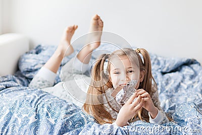 A girl with two ponytails in soft gray pajamas sits on a bed and bites a bar of chocolate in foil Stock Photo