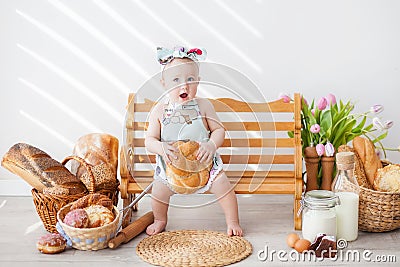 A girl up to a year old in a kitchen apron and a bow on her head sits on a bench, next to tartlets with booths Stock Photo