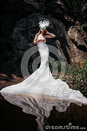 girl in a Venetian mask with feathers with a slender figure stands in the water near a rock, wrapped in a long white cloth Stock Photo