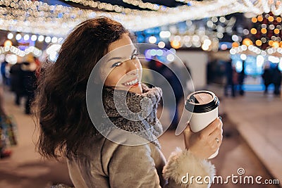 Girl Walking with a hot drink on christmas decoration lights street. Young happy smilin Woman wearing stylish knitted scarf, coat Stock Photo