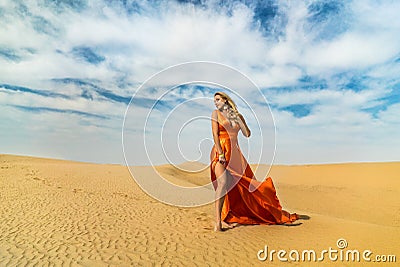 Girl walks on desert. Beautiful woman is walking, staing on sand or dune, touches, shows her legs. Blonde lady in Stock Photo