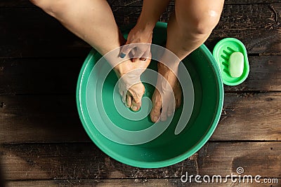 Girl washes her feet in a green bowl on the wooden floor of the house, foot care Stock Photo