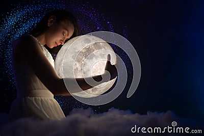 Girl in white air dress with the moon. Beautiful tender girl on the night sky with bright stars and soft clouds hugs the moon. Stock Photo