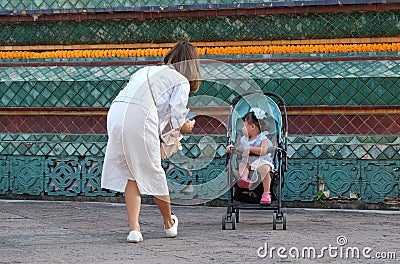 A girl in white clothes bent to take a picture of her little daughter on a cell phone. Mom Editorial Stock Photo