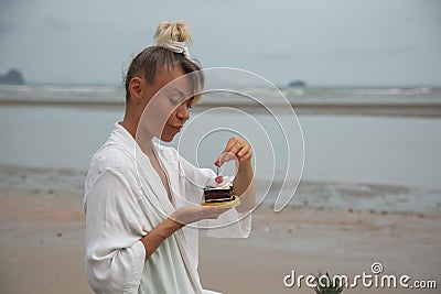Girl in white dress sitting on the beach and eating cake with cherries Stock Photo