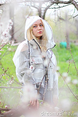 A girl with white hair, in a transparent jacket, stands near a blossoming tree with white tulips. Spring mood Stock Photo