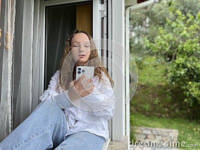 a girl in a white shirt a teenager sits on a white windowsill the view from the street she can be seen in the window in Stock Photo