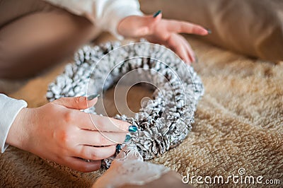 A girl in a white sweater makes a wreath of fir cones. Woman in a bedroom interior in Rustic style for Christmas and New Year. DIY Stock Photo