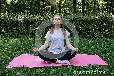 A girl in a white t-shirt and black leggings on a pink mat sits in a yoga pose on a summer green lawn Stock Photo