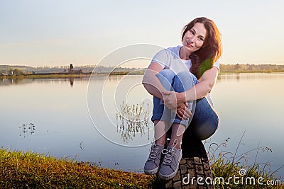 Girl or woman sitting on the shore or beach of the lake in the evening at sunset. Single trip or vacation Stock Photo